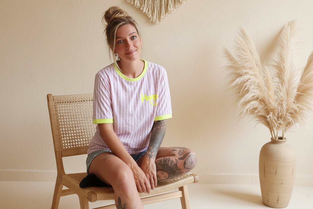 „Gestreiftes Happy T-Shirt Lila-Lavendel, Model sitzend auf Rattanstuhl, Boho-Luxe Setting – SK-Style