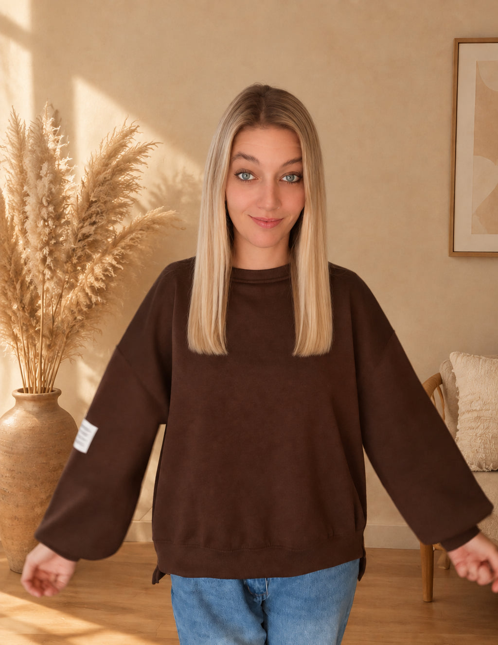 Vorderansicht des dunkelbraunen Damen-Sweatshirts mit hellem Ärmelpatch, am Model mit leicht ausgestreckten Armen und vollständig sichtbarem Saum.