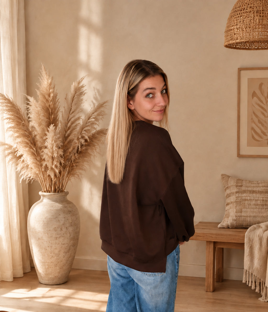 Seitliche Rückansicht des dunkelbraunen Damen-Sweatshirts mit lockerem Schnitt, am Model zur blauen Jeans mit Blick über die Schulter.