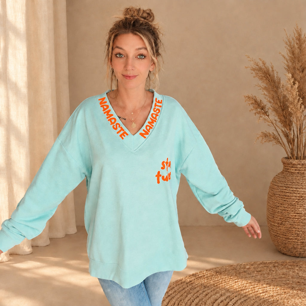 Damenpullover in Mint mit orangefarbenem Namasté-Schriftzug am V-Ausschnitt und Stay-tuned-Print, Frontansicht am Model