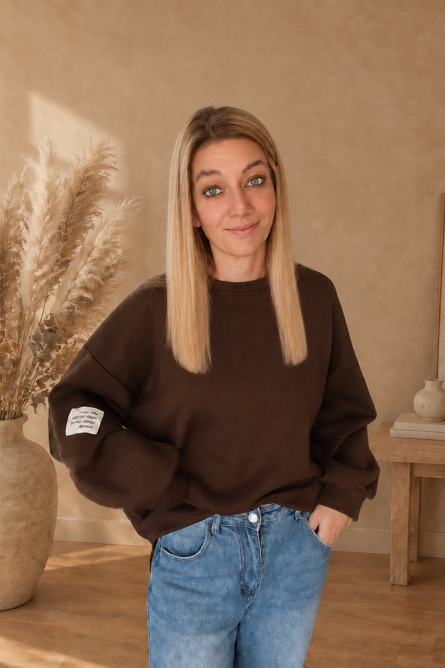 Nähere Vorderansicht des dunkelbraunen Damen-Sweatshirts mit hellem Statement-Patch, vorne in eine blaue Jeans gesteckt, eine Hand in der Jeanstasche.