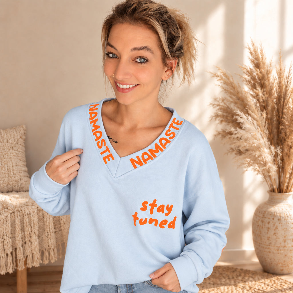 Detailansicht des hellblauen Pullovers mit orangefarbenem Namaste-Schriftzug am V-Ausschnitt und „stay tuned“-Print