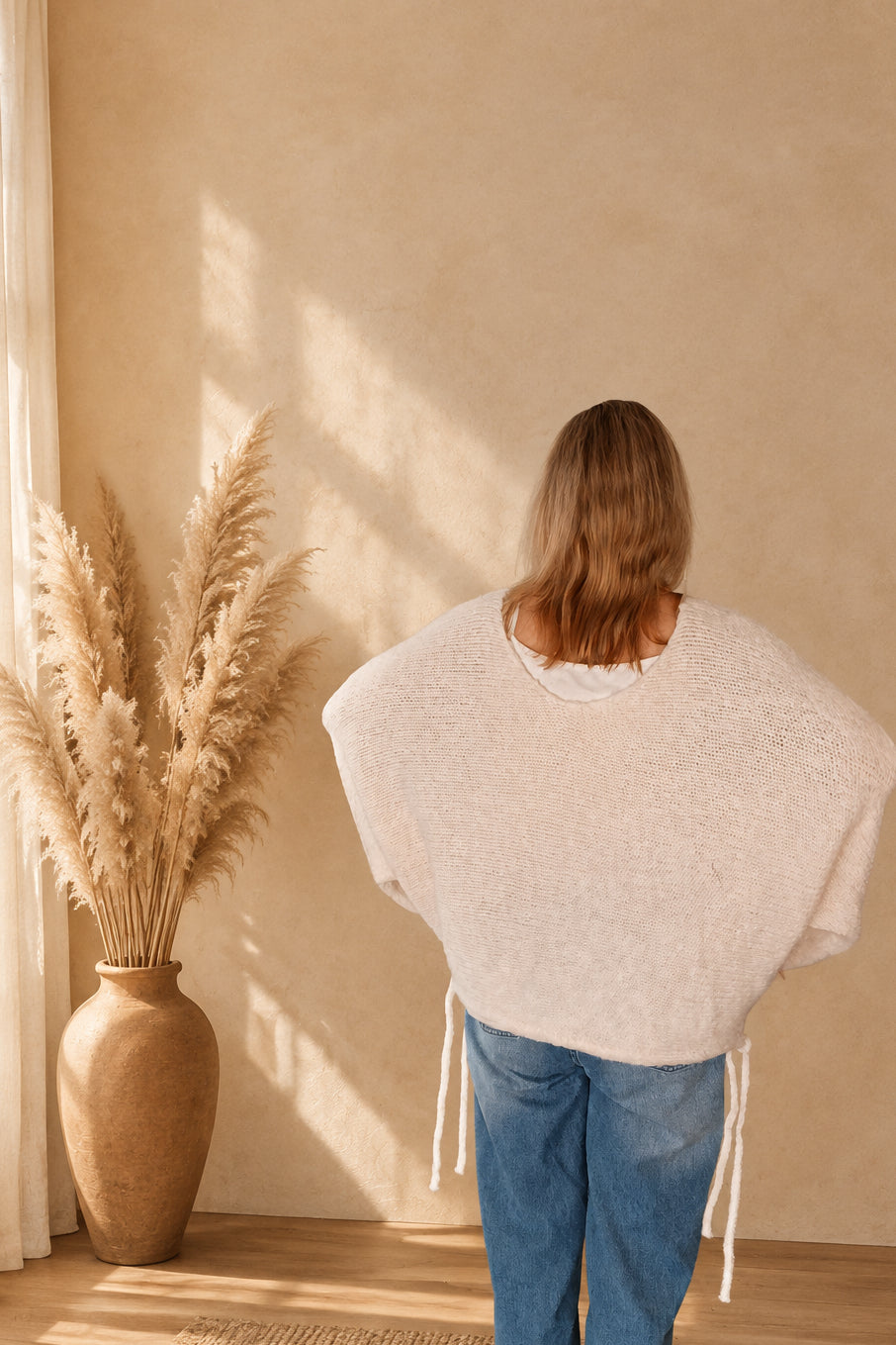 Cremeweißer Oversized-Sweater von hinten am Model, mit weit geschnittenen Ärmeln und seitlichen Bändern.