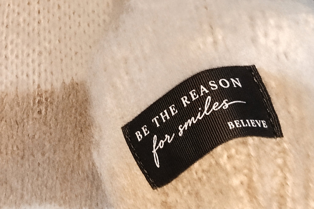 Nahaufnahme des schwarzen Ärmelpatches mit Schriftzug „BE THE REASON for smiles BELIEVE“ auf beige-cremefarbenem Strick
