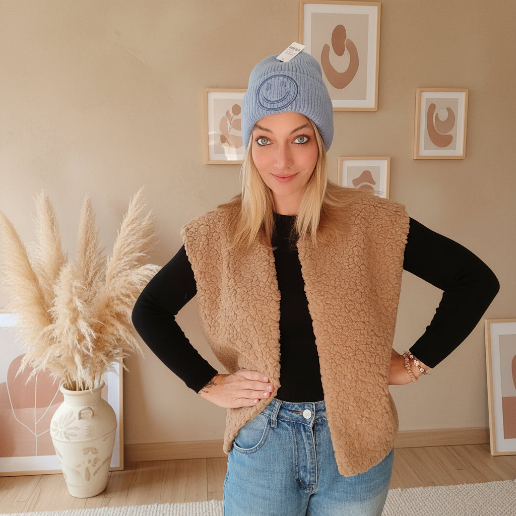 Hellblaue Smiley-Beanie in frontaler Outfitansicht, getragen zu schwarzem Oberteil, beiger Weste und Jeans