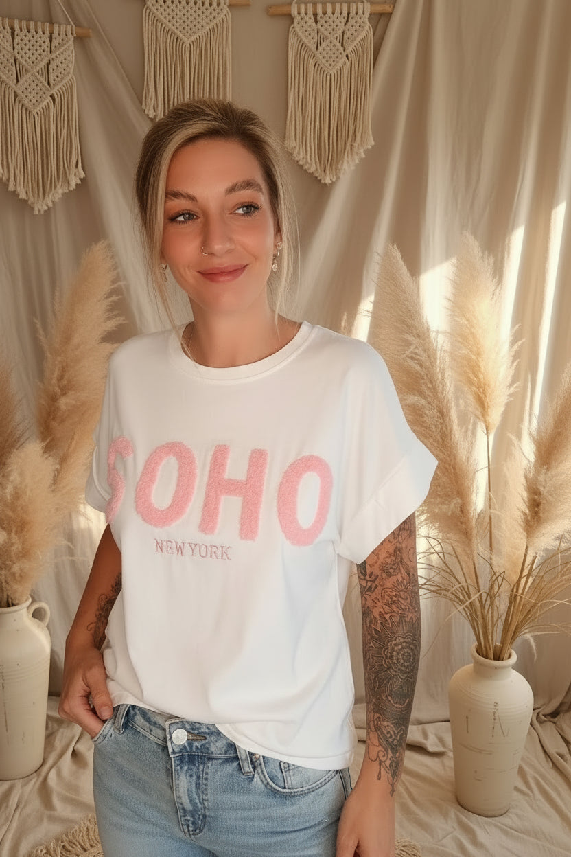 Boho-Luxe weißes Baumwoll-T-Shirt mit rosa SOHO-Schriftzug vor Makramee-Deko.