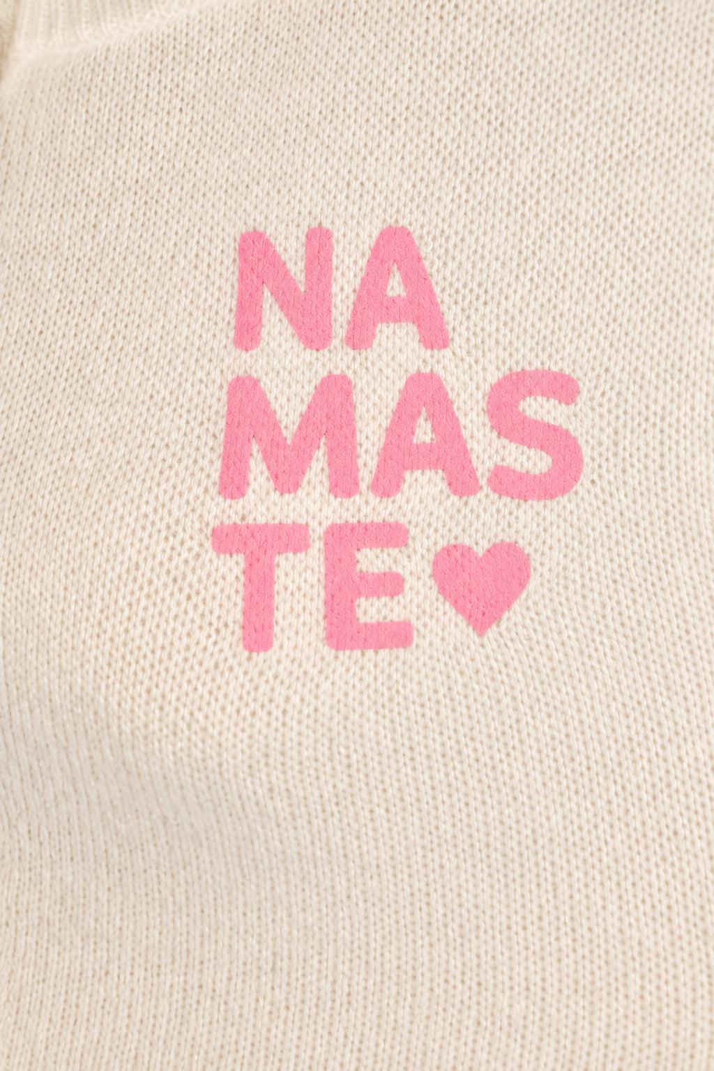 Detail des rosa Namasté-Schriftzugs mit Herz auf cremeweißem Strickpullover.