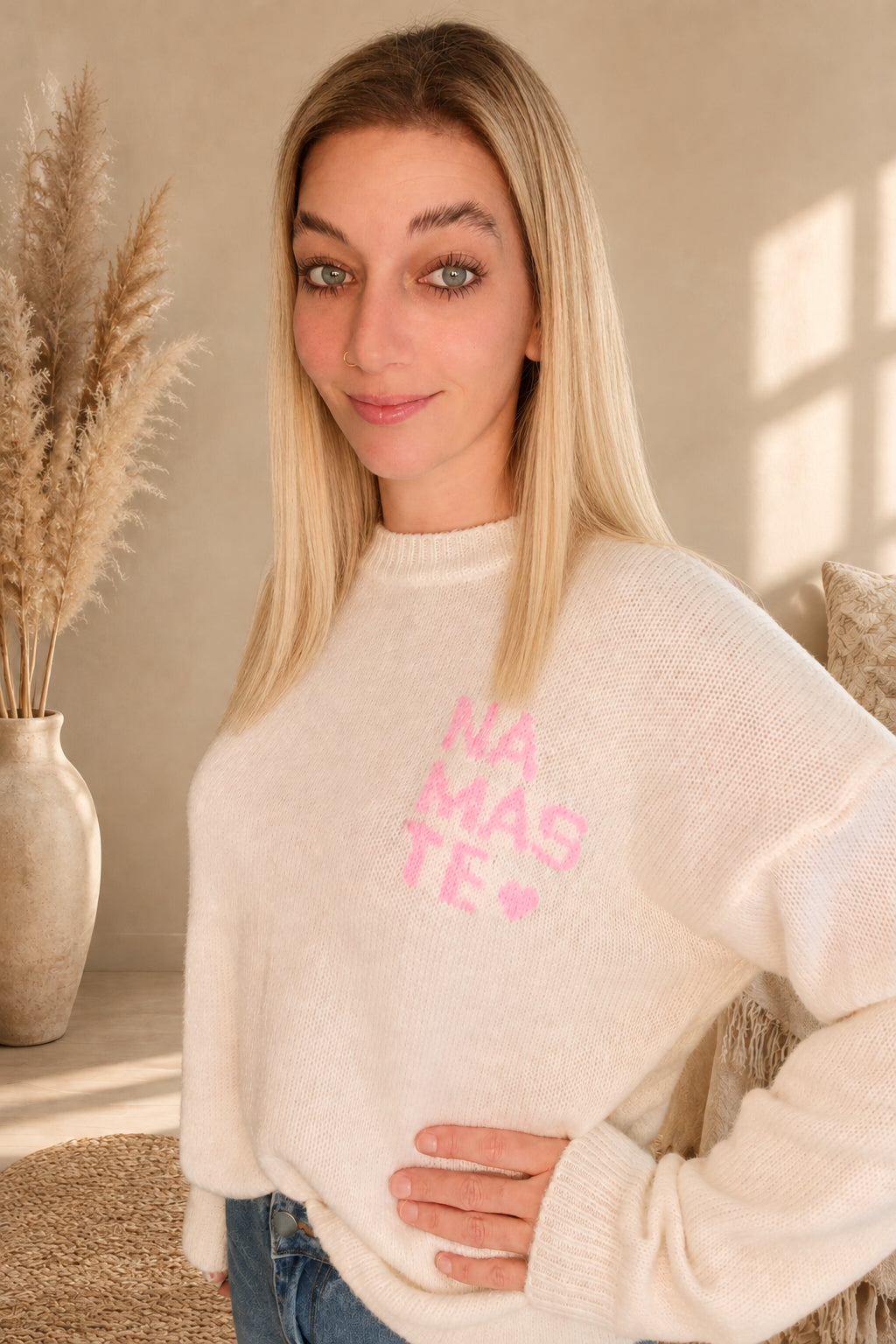 Detailansicht des cremeweißen Namasté-Strickpullovers mit rosa Brustprint, am Model mit Hand an der Hüfte.