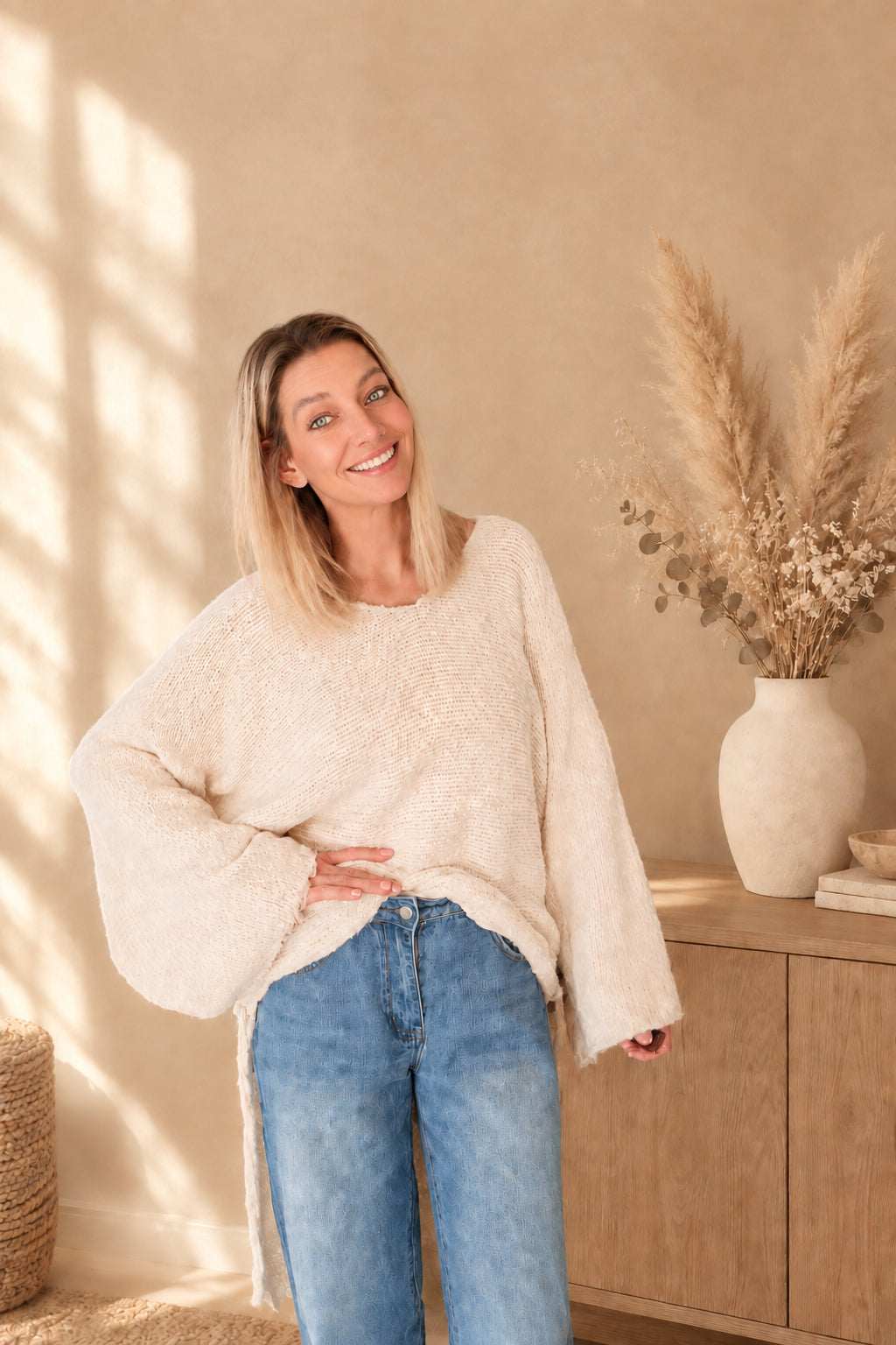 Cremeweißer Oversized-Sweater locker zu blauen Jeans gestylt, mit weitem Ärmel und sichtbarem seitlichem Band.