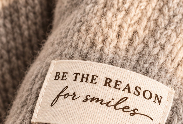 Nahaufnahme des hellen Patches „Be the reason for smiles“ auf der grauen Strickstruktur des Damenpullovers.