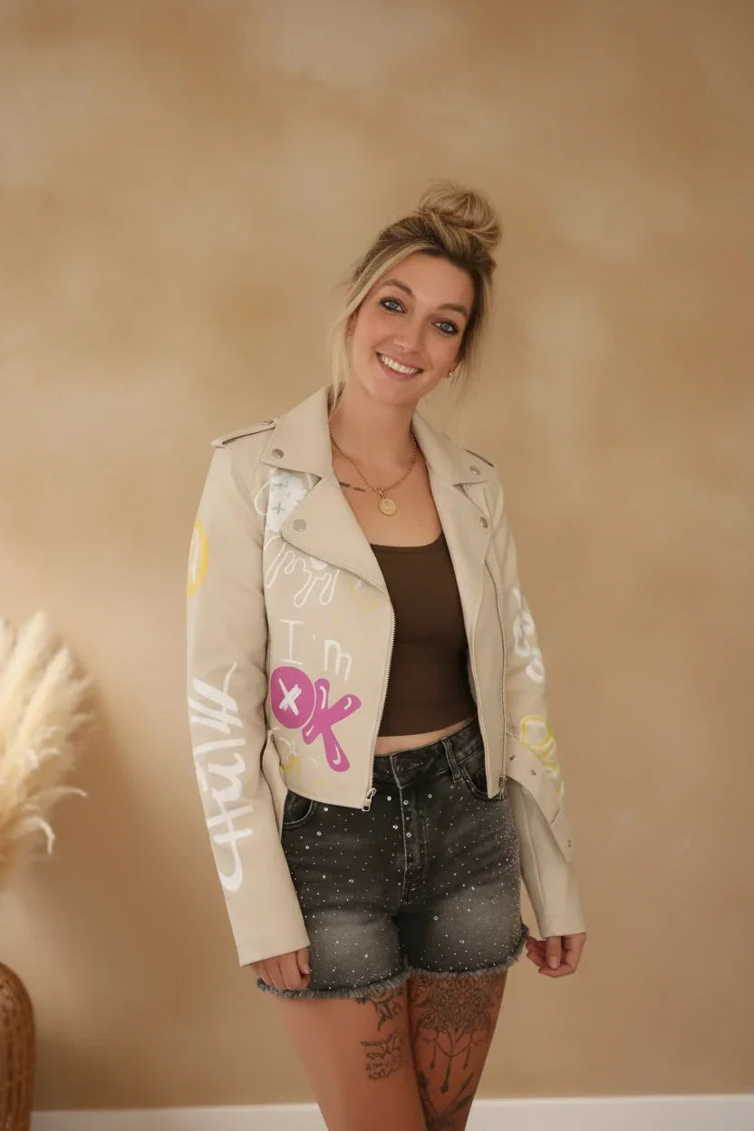 Frau in beiger Lederjacke mit Graffiti-Prints, braunem Top und glitzernden Denim-Shorts