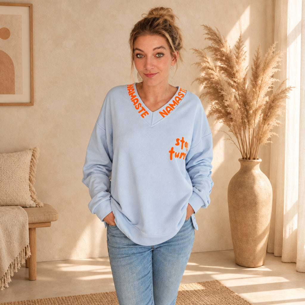 Hellblauer Damenpullover mit V-Ausschnitt und orangefarbenen Prints, Frontansicht mit Händen in den Hosentaschen