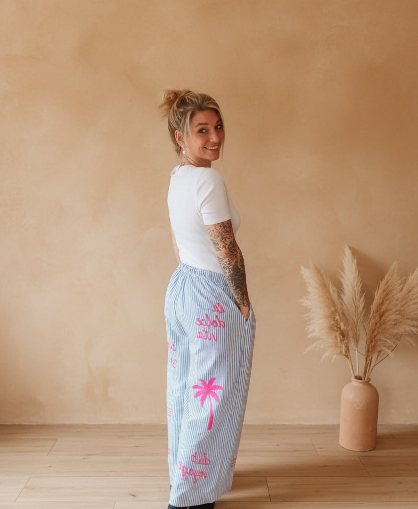 Boho-Luxe Look: Frau in weißem T-Shirt und hellblauen, gestreiften Baumwollhosen mit Palmen-Print.