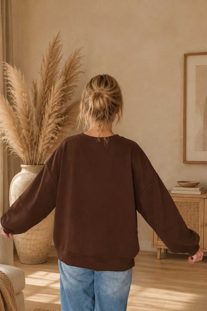 Rückansicht des dunkelbraunen Damen-Sweatshirts mit überschnittenen Schultern und weiten Ärmeln, am Model mit Dutt und leicht ausgestreckten Armen.