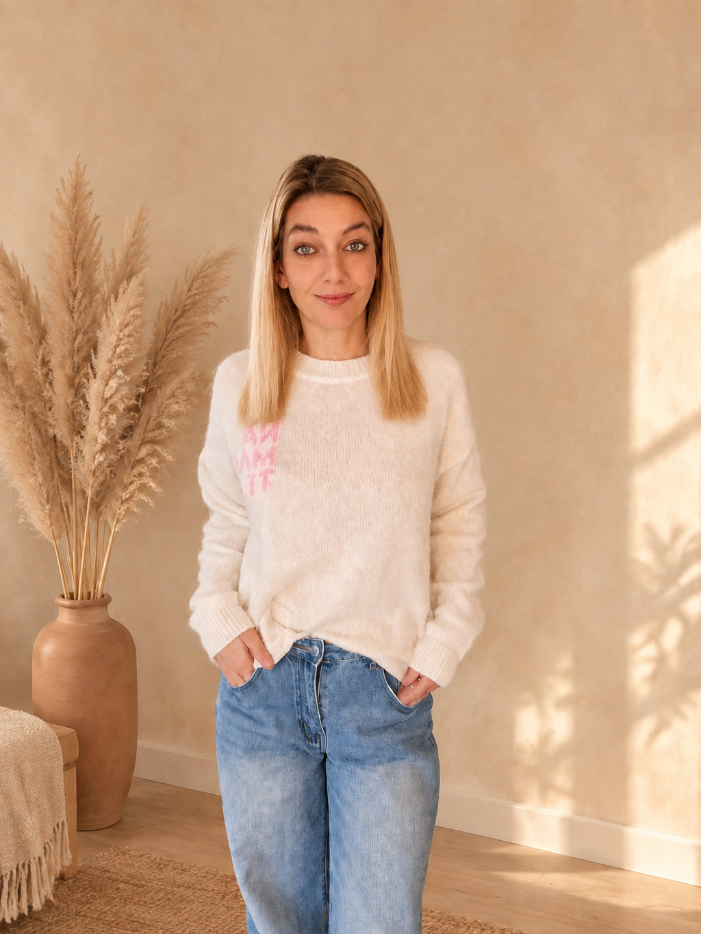 Cremeweißer Namasté-Strickpullover mit rosa Brustprint, vorne teilweise in eine blaue Jeans gesteckt.