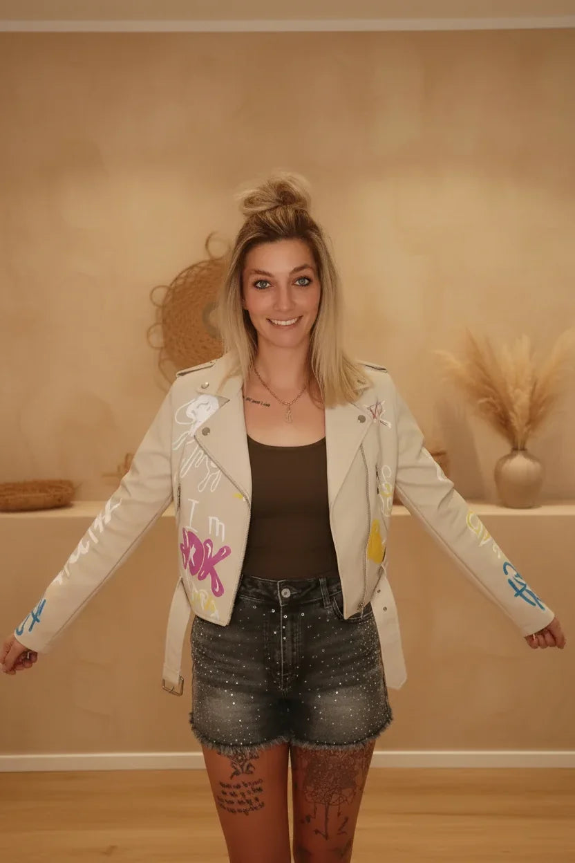 Frau in beiger Kunstleder-Jacke mit Graffiti-Prints, braunem Top und glitzernden Denim-Shorts.