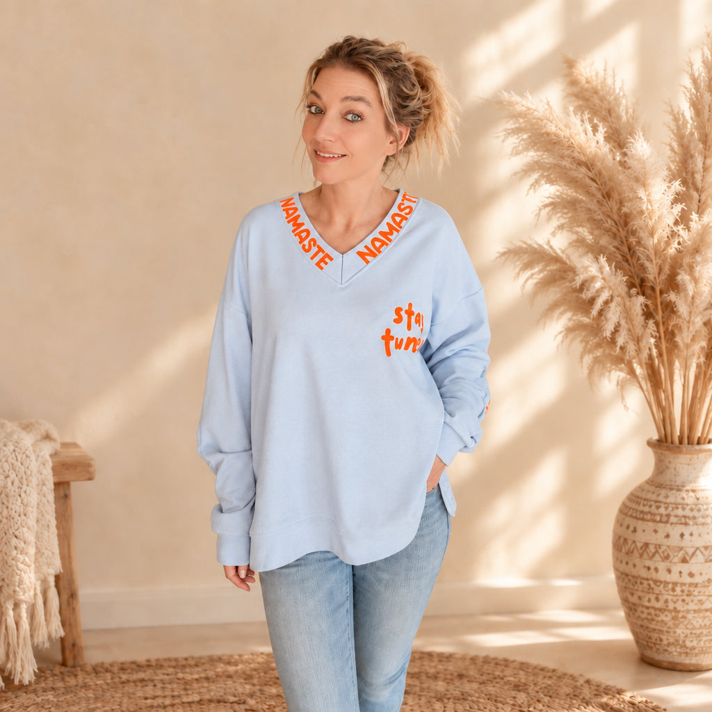 Hellblauer Namaste-Damenpullover mit orangefarbenem Schriftzug am V-Ausschnitt und „stay tuned“-Print, leicht seitliche Frontansicht
