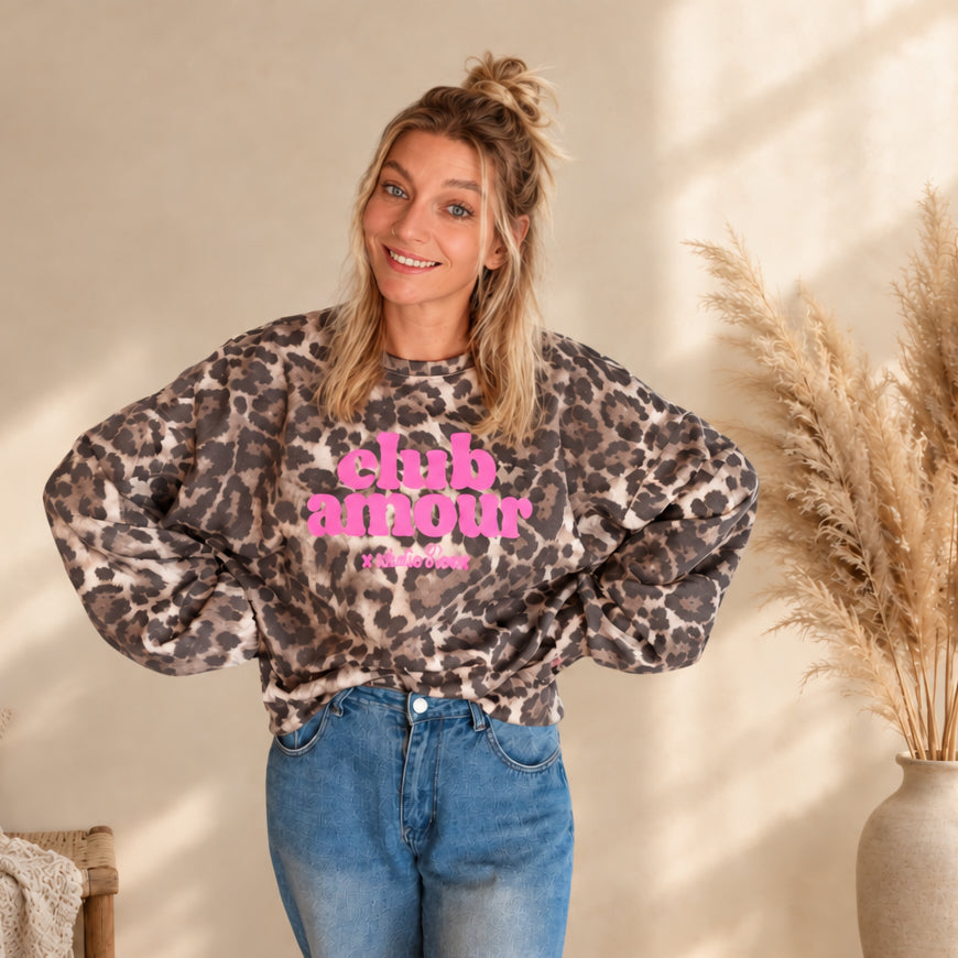 Pullover mit Leo-Print in Beige und Braun und pinkem „club amour“-Schriftzug, frontal am Model mit den Händen an der Taille.