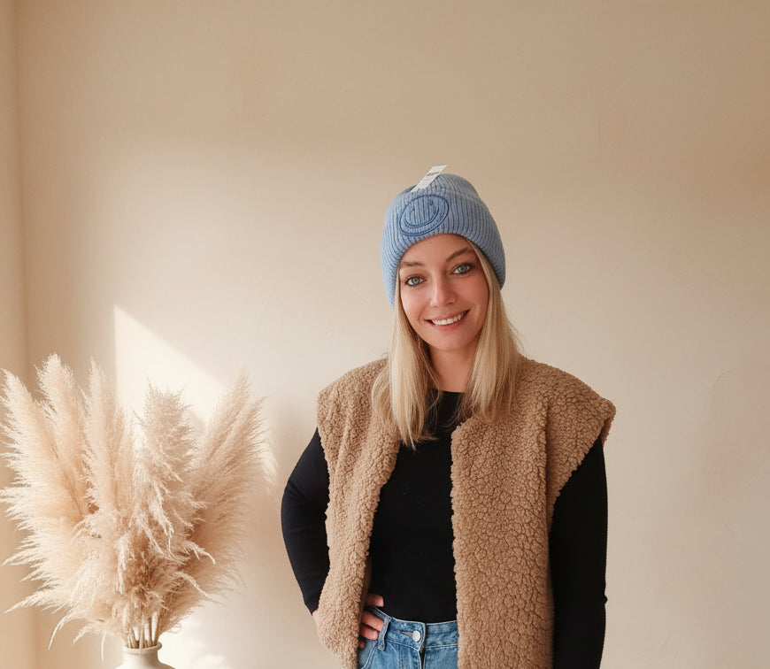 Hellblaue Smiley-Mütze als Beanie an einer Frau getragen, frontale Outfitansicht mit beiger Weste und Jeans